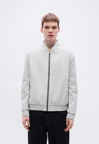 Ανοιχτό γκρι μπουφάν bomber με μπροστινό φερμουάρ, ριμπ μανσέτες και λεία υφή. Στυλιζαρισμένο με σκούρα τζιν, με μινιμαλιστικό σχεδιασμό.