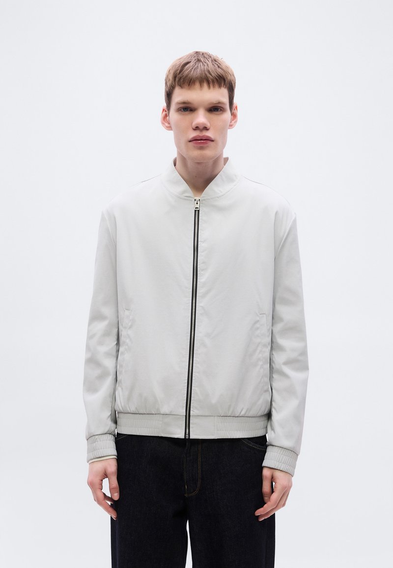 Ανοιχτό γκρι μπουφάν bomber με μπροστινό φερμουάρ, ριμπ μανσέτες και λεία υφή. Στυλιζαρισμένο με σκούρα τζιν, με μινιμαλιστικό σχεδιασμό.