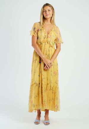 Młoda kobieta stoi ze złączonymi rękami, ubrana w żółtą, kwiatową sukienkę maxi z falbanami na rękawach i jasnoniebieskie sandały na obcasie z odkrytymi palcami.