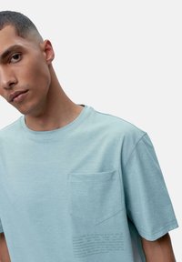 Šviesiai mėlynas, trumpomis rankovėmis marškinėliai iš minkštos medžiagos, su kairėje krūtinės pusėje esančia kišene ir tekstu apatiniame šone. Klasikinis apvalios apykaklės dizainas.