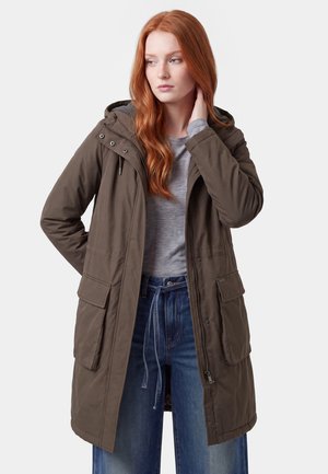 Junge Frau mit roten Haaren trägt einen braunen Kapuzenmantel über einem grauen Shirt und blauen Jeans, schaut zur Seite mit einer Hand nahe am Hals.
