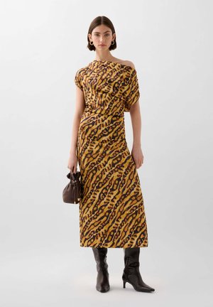 Geel en bruin dierenprint jurk met een off-shoulder ontwerp, gecombineerd met kniehoge zwarte laarzen en een kleine donkere handtas.
