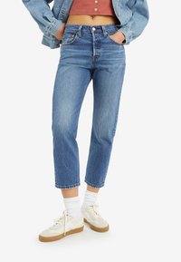 Personne portant un jean bleu court, des chaussettes blanches, des baskets beiges, un haut court rouille avec boutons, et une veste en jean bleue, les mains dans les poches.