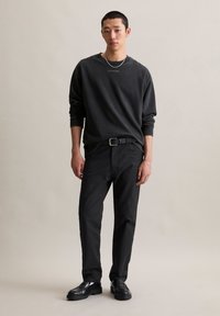 Jonge man in een zwart langslavend shirt, zwarte broek met riem, zwarte schoenen, zilveren ketting en armband die tegen een effen achtergrond staat.