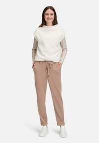 Biały bezrękawnik z polaru na paskowanej koszuli z długim rękawem, zestawiony z beżowymi zwężanymi spodniami i białymi tenisówkami. Detal ze ściągaczem w talii.