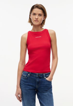 Kvinde iført ærmeløs rød tanktop med "tommy jeans"-logo og blå jeans, stående med den ene hånd i lommen mod hvid baggrund.