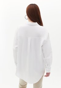 Camicia bianca a maniche lunghe con vestibilità rilassata, piega posteriore e orlo arrotondato, realizzata in un tessuto morbido, abbinata a pantaloni beige.