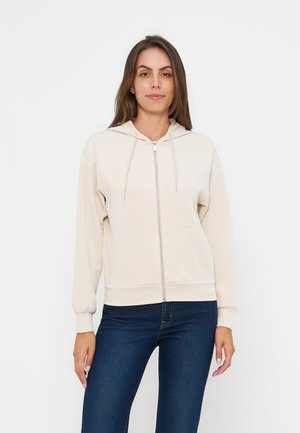 Donna con felpa con cappuccio beige chiaro con zip e tasche frontali e jeans blu scuro, in piedi davanti a uno sfondo bianco uniforme.