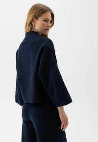 Giacca cropped blu navy in tessuto morbido, con colletto a rever e ampie maniche a tre quarti, abbinata a pantaloni scuri coordinati.