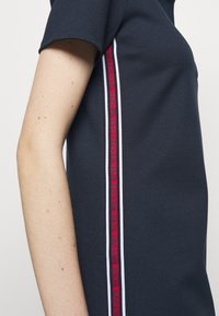 Mörkblå polo skjorta med texturerad tyg, korta ärmar och ett rödvitt randig detalj längs sidan med varumärkestext.