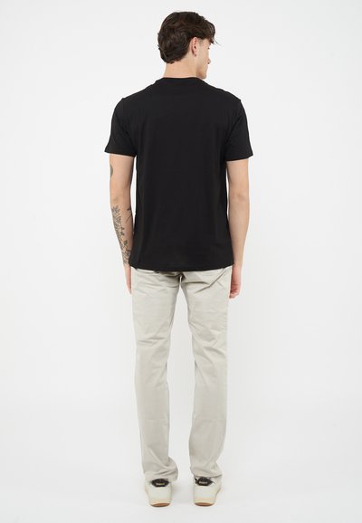 Hombre vestido con una camiseta negra de manga corta, pantalones beige y zapatillas deportivas blancas, de pie de espaldas sobre un fondo blanco liso.