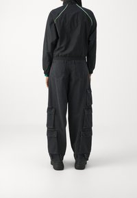 Svart vindjacka med gröna detaljer, kombinerad med löst sittande svarta cargobyxor med flera fickor och en texturerad yta.