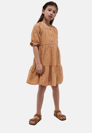 Jeune fille debout portant une robe fleurie pêche avec boutons et des sandales marron sur un fond clair uni.