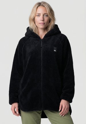 Frau trägt eine schwarze Fleecejacke mit Reißverschluss und Kapuze sowie grüne Hose, steht vor einem schlichten hellen Hintergrund.