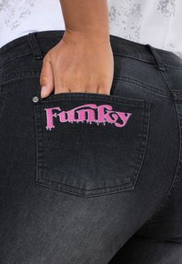 Černé džíny z denimu s zadní kapsou, na které je růžový nápis "Funky". Ruka zasunutá do kapsy ukazuje švy a detaily kování.