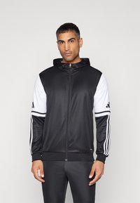 Felpa con zip bianco e nero, dotata di cappuccio, cerniera frontale e tre strisce sulle maniche. Realizzata in tessuto liscio e leggero.