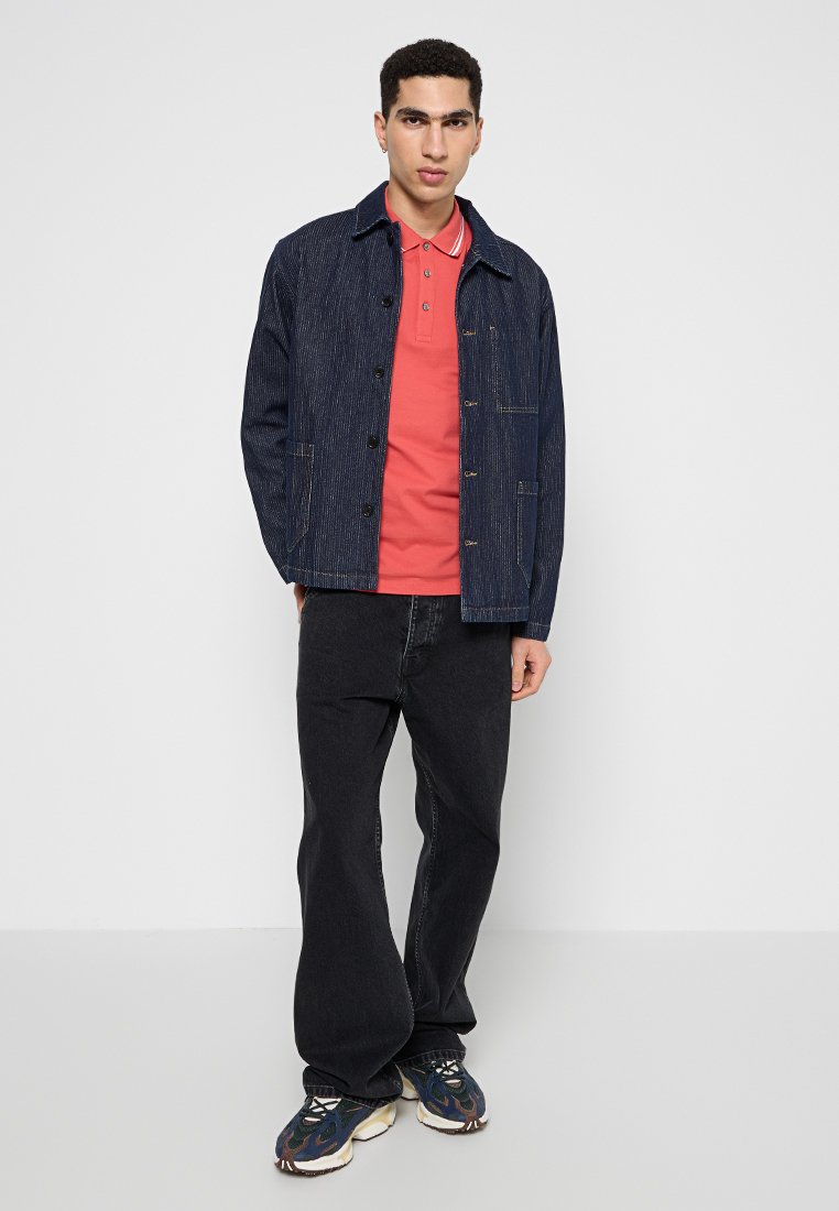 Ciemnoniebieska kurtka dżinsowa na czerwonym polo, w połączeniu z szerokimi, czarnymi spodniami dżinsowymi i wzorzystymi sneakersami. Skupienie na stylu casual streetwear.