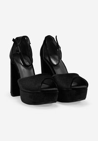 Tacones de plataforma negros con una textura de terciopelo suave, con punta abierta y correa al tobillo con hebilla. El alto tacón de bloque proporciona una elevación significativa.