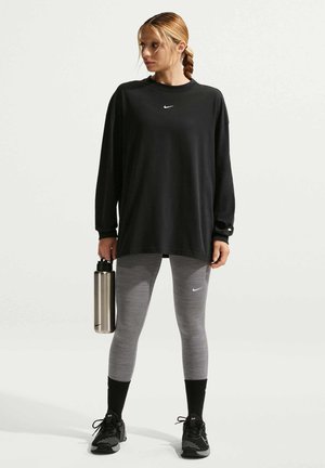 Mujer con camiseta negra de manga larga y leggings grises sosteniendo una botella de agua plateada, de pie contra un fondo claro y sencillo.