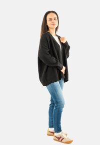 Mujer de pie con un atuendo casual que incluye un cárdigan negro extragrande, una camiseta blanca, jeans azules y zapatillas beige con rayas burdeos, sobre un fondo blanco.