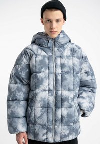 Szara kurtka puchowa tie-dye z kapturem, z pikowanym wzorem i srebrnym zamkiem. Miękka tekstura i luźny krój. Na głowie czarna czapka.
