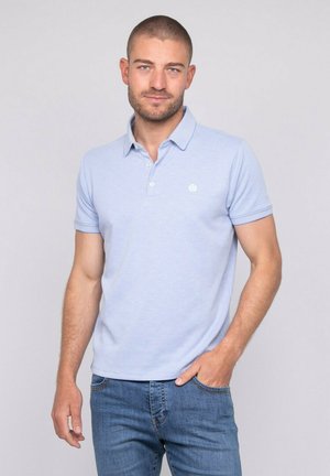 Homme aux cheveux courts et à la barbe, portant un polo bleu clair et un jean bleu, debout avec une main dans la poche devant un fond uni.