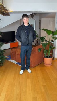 Jonge man in een donker jack, spijkerbroek en witte sneakers staand in een woonkamer met een grote potplant en een oranje bank.