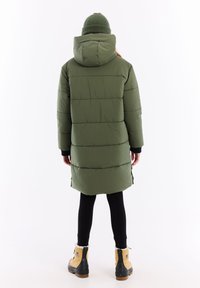 Lang, puffer, olijfgroene jas met een capuchon, voorzien van horizontale stiksels en zijsplitten, gecombineerd met zwarte leggings en tan kleurige laarzen.