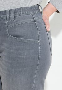 Jeans de mezclilla gris con cintura elástica, que presentan bolsillos cosidos y una textura suave, mostrados de lado con una mano en el bolsillo.