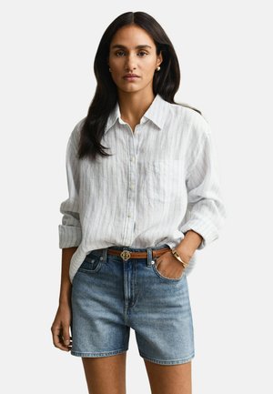 Femme aux longs cheveux foncés portant une chemise blanche à boutons avec les manches retroussées et un short en denim bleu avec une ceinture marron.