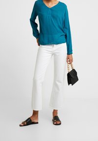 Blouse à manches longues bleu sarcelle avec un col en V, associée à un jean blanc évasé. Sandales plates noires et petit sac à main noir avec poignées en bois.