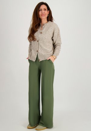 Vrouw met lang bruin haar, gekleed in een beige vest met knopen, olijfgroene broek met wijde pijpen en beige schoenen, staand met handen in de zakken.