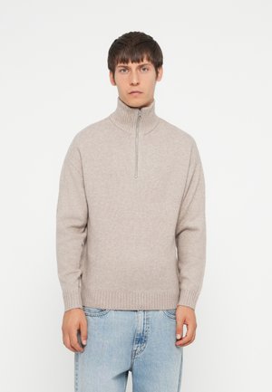 Light beige knitted sweater with a high collar and half-zip closure. Features ribbed cuffs and hem, worn over light blue jeans.