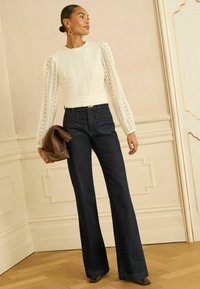 Vit långärmad topp i spets med strukturerade ärmar, kombinerad med mörkblå utsvängda jeans. Modellen håller en brun clutch och står på ett mönstrat golv.