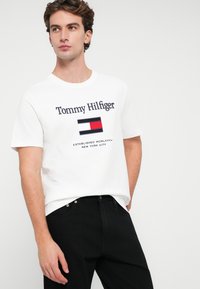 Hvid bomulds T-shirt med "Tommy Hilfiger" logo, et marineblåt rektangel mønster med røde og hvide detaljer. Båret med sorte jeans.