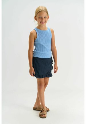 Smilende pige stående iført en blå ærmeløs ribbet top, marineblå nederdel med flæser og beige sandaler mod en ensartet hvid baggrund.