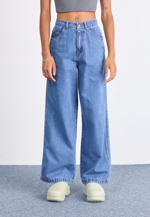 Personne portant un jean bleu taille haute à jambes larges, un top court gris, des chaussures beiges à plateforme et un anneau de nombril argenté, debout sur un tapis.