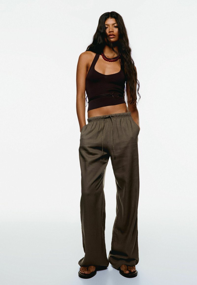 Tmavohnedý crop top s halter krkom; voľné olivovo zelené nohavice s elastickým pásom; nosené s čiernymi sandálmi.