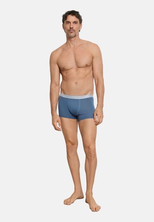 Erwachsener Mann steht barfuß, trägt blaue Boxer-Shorts mit einem grauen Bund, vor einem schlichten weißen Hintergrund.