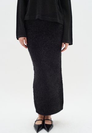 Persoon draagt een lange zwarte pluizige rok, zwarte longsleeve top en zwarte puntige platte schoenen met bandje, staand op een witte vloer.
