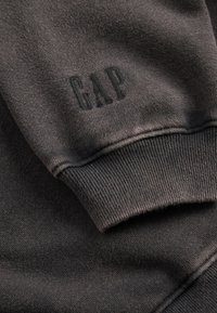 Tmavě šedá mikina s žebrovanými manžetami. Na rukávu je jemně vyražené logo „GAP“. Měkká textilie, pohodlný design.