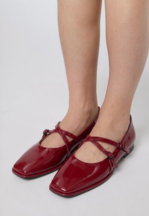 Fødder iført skinnende røde Mary Jane-flade sko med firkantede tåsnuder og krydsede spænder mod en ensfarvet lys baggrund.