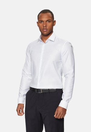 Camisa blanca de manga larga con un patrón sutil, cuello tipo abotonado y detalles en los puños. Llevada con pantalones oscuros y un cinturón negro.