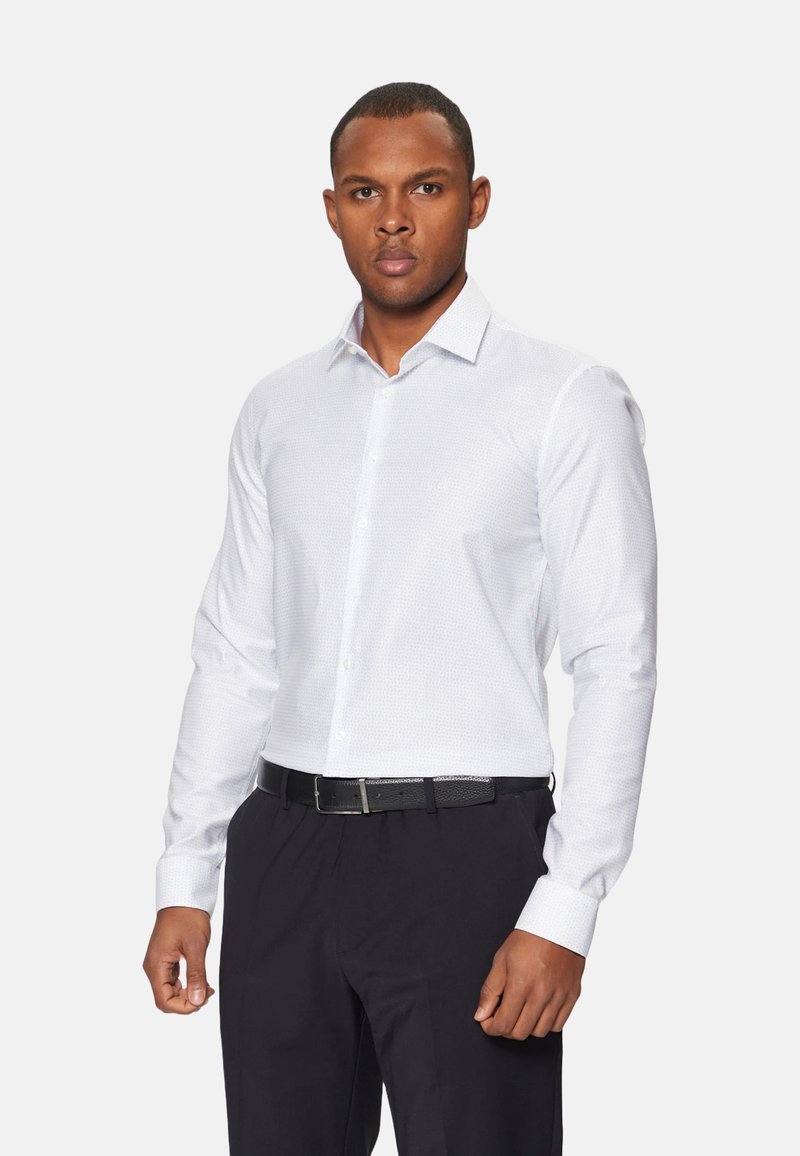 Camisa blanca de manga larga con un patrón sutil, cuello tipo abotonado y detalles en los puños. Llevada con pantalones oscuros y un cinturón negro.