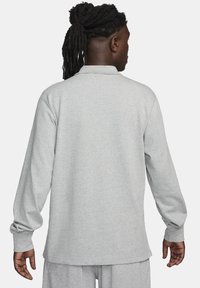 Harmaa pitkähihaisen poolopaita, joka on valmistettu pehmeästä kankaasta. Paidan leikkauksessa on klassinen kaulus ja rento istuvuus, ja helmassa on näkyviä ompelutöitä.