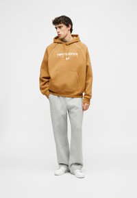 Νέος άνδρας όρθιος, φοράει καφέ φούτερ New Balance, ανοιχτόγκρι φόρμα και λευκά αθλητικά παπούτσια, κοιτώντας στο πλάι.