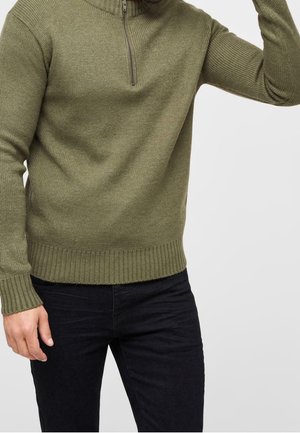 Mężczyzna ubrany w oliwkowo zielony dzianinowy sweter z półzamkiem, do tego czarne spodnie, na tle jednolitego jasnoszarego tła.