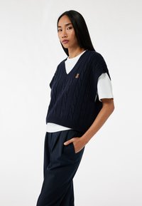 Marineblå strikket vest med kabelmønster og V-halsdesign, med et broderet bjørnelogo på venstre bryst. Båret med en hvid skjorte under.