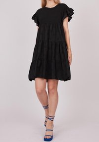 Robe noire à volants avec des manches courtes en dentelle, présentant une silhouette fluide. Portée avec des sandales à talons bleues pour un contraste. Texture de tissu lisse.