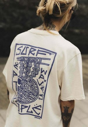 Osoba o blond włosach związanych w kok, ubrana w beżowy T-shirt z niebieskim abstrakcyjnym wzorem i napisem "SURF WAVE SIMPLE" na plecach.